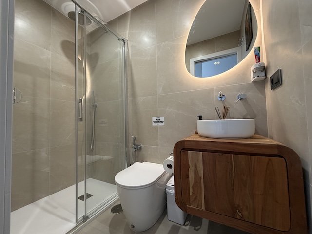 Baño pequeño con ducha de mampara, lavabo sobre mueble de madera y espejo redondo retroiluminado.