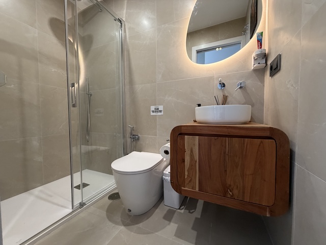 Vista del baño pequeño desde la entrada con lavabo, espejo redondo retroiluminado y paredes en tono piedra.