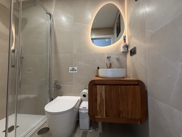 Lavabo sobre mueble de madera con grifería mural y espejo retroiluminado en pared tono piedra.