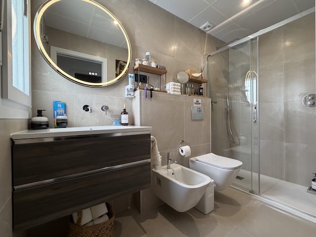 Baño principal con lavabo, espejo retroiluminado, bidé, WC y ducha con mampara de cristal.