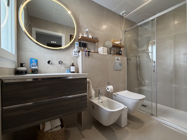 Vista del baño principal con mueble lavabo, espejo redondo con luz y ducha con mampara.