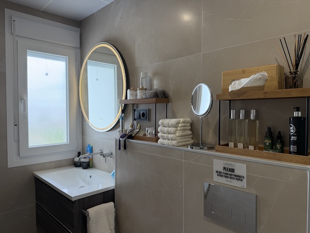 Vista general del baño principal con lavabo, espejo retroiluminado, estanterías con amenities y ventana.