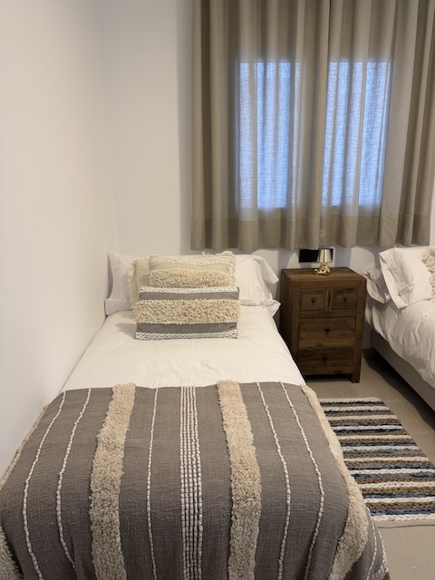 Cama individual con cojines decorativos y manta en tonos beige y gris.