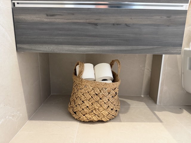 Cesta de fibras naturales con rollos de papel higiénico bajo el mueble del lavabo.