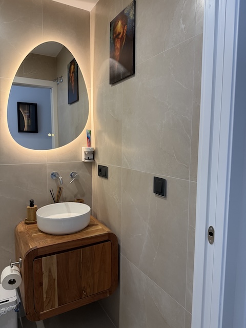 Detalle de lavabo circular blanco sobre mueble de madera con grifería mural en baño pequeño.
