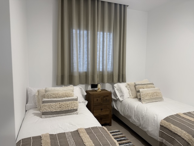 Dormitorio con dos camas individuales, mesita de madera y cortinas en tonos neutros.