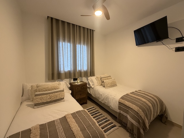 Vista general del dormitorio con dos camas, alfombra a rayas y TV en pared.