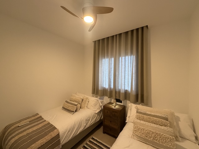 Dormitorio con dos camas individuales, ventilador de techo, mesita de madera y cortinas en tonos neutros.