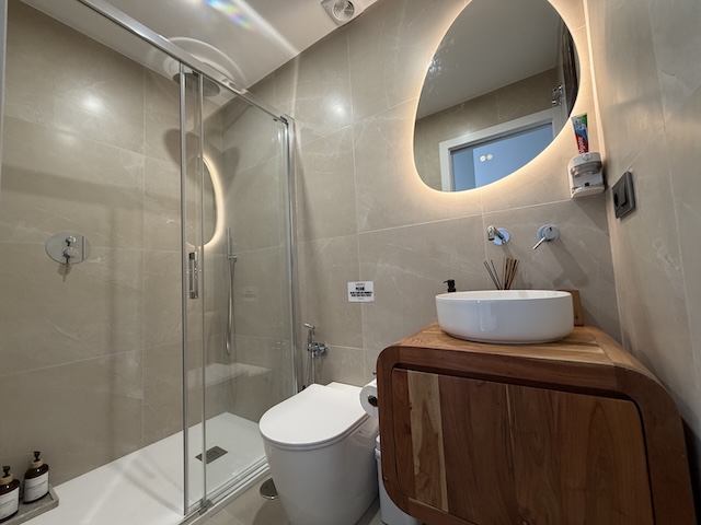 Vista general del baño pequeño con ducha de mampara, inodoro moderno y lavabo sobre mueble de madera.